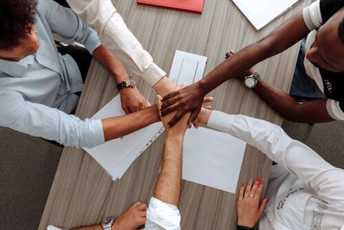 A top-down view of a diverse group of people around a table, stacking their hands together in a gesture of unity and teamwork.