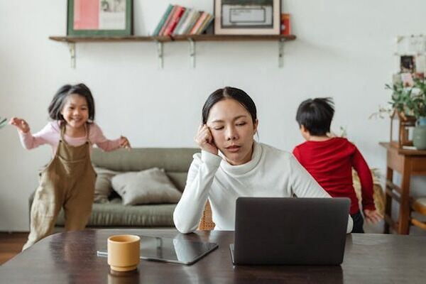 Stressed mom looking at a laptop screen while the kids play in the background. 