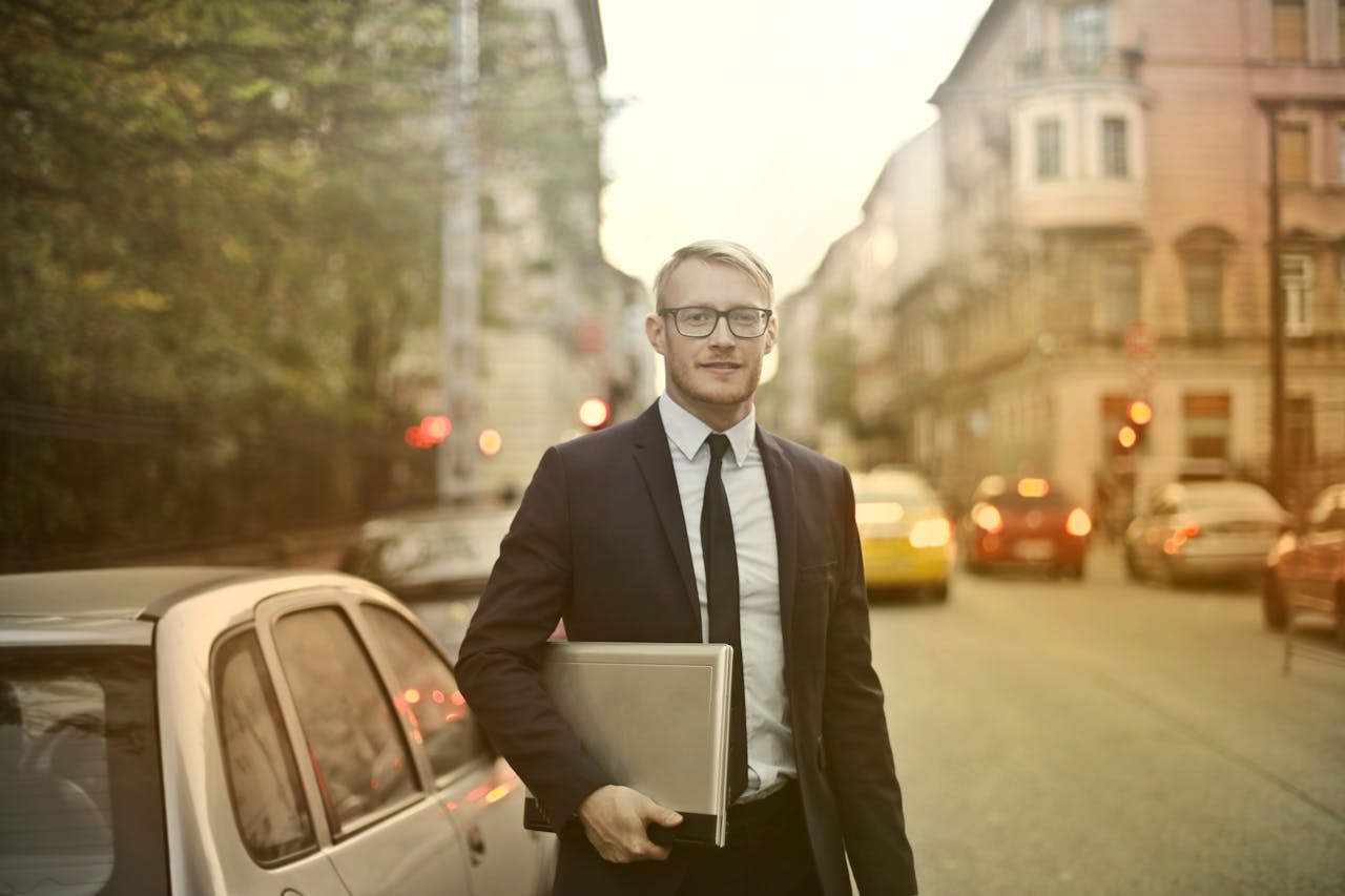 A businessman with a laptop stands near a car on the street.