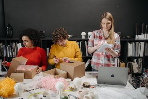 People putting things in cardboard boxes, labeling them, with a computer and shelves.