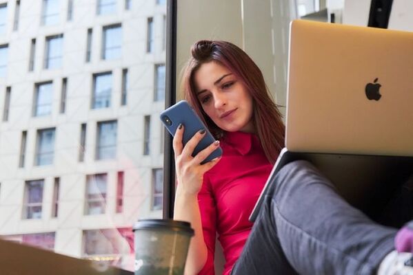 A woman in a red shirt works remotely, looking at her phone while sitting with a laptop and coffee.