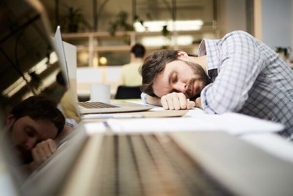 A man sleeping at his desk, resting his head on his arm with an open laptop and papers around him.