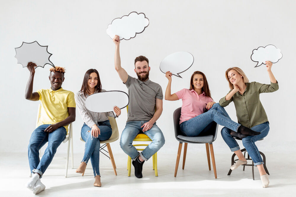 A group of five diverse people sitting on chairs, smiling and holding empty speech bubble boards above their heads.