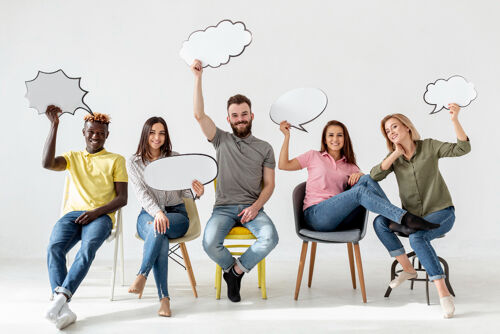 A group of five diverse people sitting on chairs, smiling and holding empty speech bubble boards above their heads.