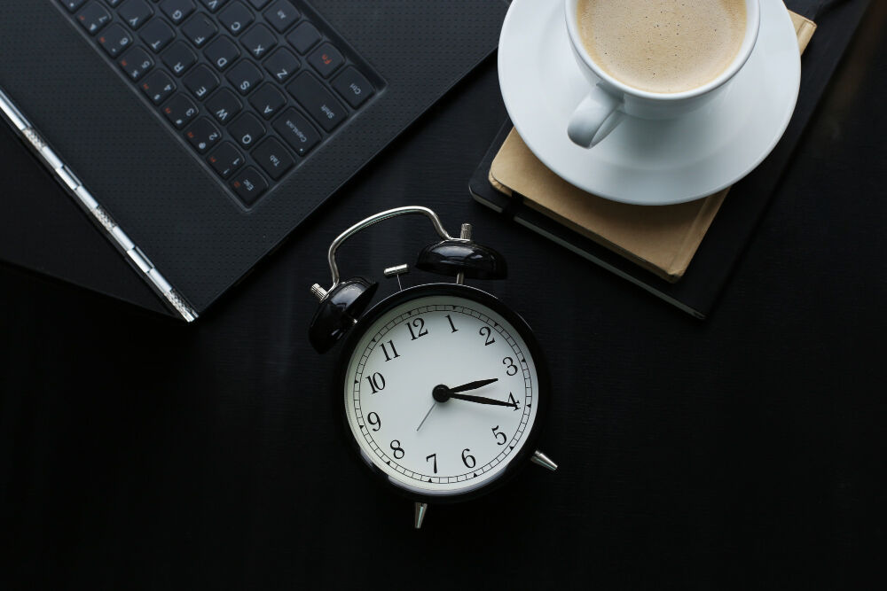An alarm clock on a desk with a notebook, pen, laptop keyboard, and a cup of coffee indicates time management for productivity.